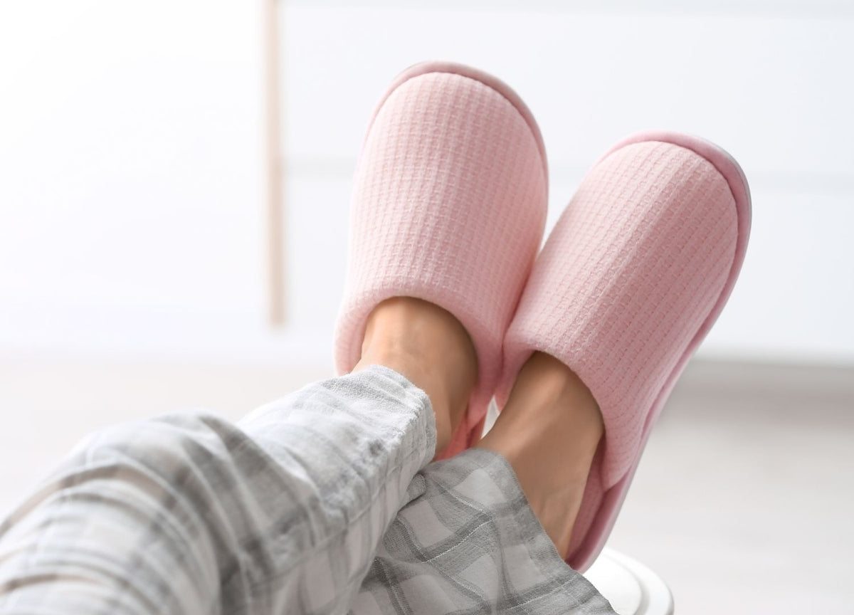 Someone wearing pink orthopedic slippers and grey pyjama pants.