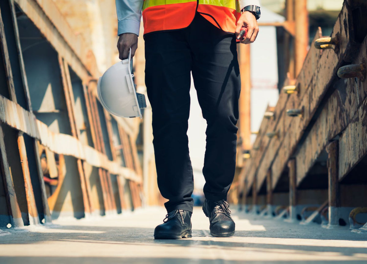 A man in construction vest and hard hat wearing work boots walking on a site. 